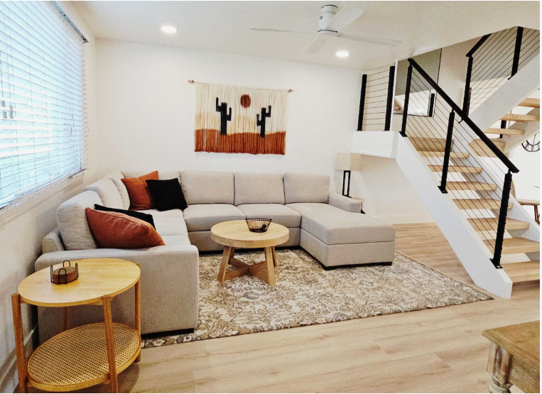 Small Living Room Decor Ideas on a Budget: 25 Stylish Ways to Make It Look Expensive Cozy living room featuring small living room decor ideas on a budget with a light beige sectional sofa, a round wooden coffee table, a matching side table, a patterned area rug, a desert-themed wall hanging, and stairs with modern black and cable railings.