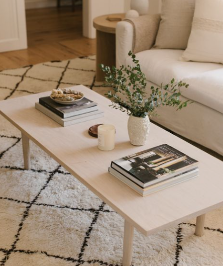 Small Living Room Decor Ideas on a Budget: 25 Stylish Ways to Make It Look Expensive Cozy living room featuring small living room decor ideas on a budget with a light wood coffee table styled with stacked books, a small vase of greenery, and a candle, all resting on a patterned white and black area rug next to a white sofa.