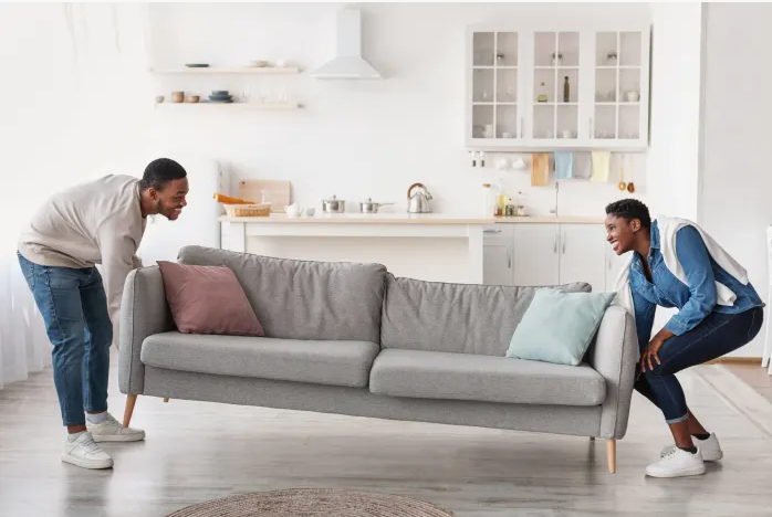 Small Living Room Decor Ideas on a Budget: 25 Stylish Ways to Make It Look Expensive A smiling couple lifting a light gray sofa with pink and blue accent pillows in an open-concept living and kitchen area featuring small living room decor ideas on a budget.