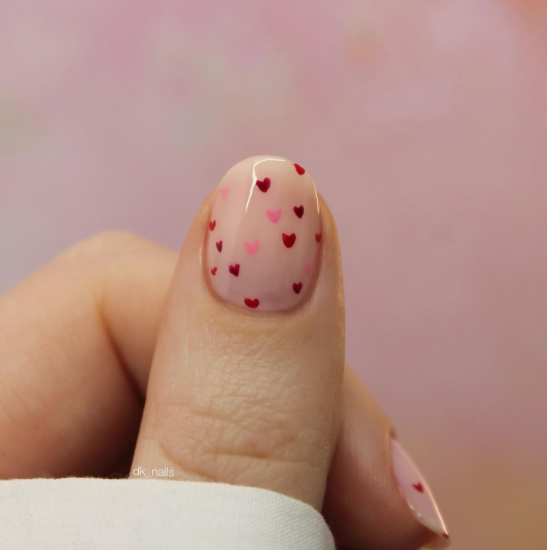 Short nail designs featuring tiny, scattered red and pink heart motifs on a neutral, glossy base.