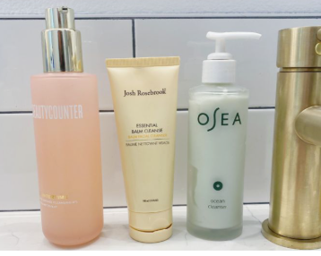 Line-up of premium beauty products worth buying featuring various facial cleansers from brands like Beautycounter, Josh Rosebrook, and OSEA arranged next to a modern faucet.