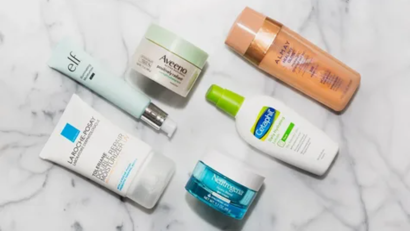 Flat lay of drugstore beauty products worth buying featuring an assortment of affordable moisturizers and lotions from brands like e.l.f., Aveeno, Almay, La Roche-Posay, Cetaphil, and Neutrogena arranged on a marble tabletop.