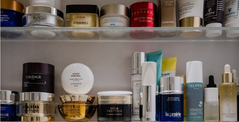 An organized collection of luxury skincare products worth buying featuring various high-end creams, serums, and jars from prestigious brands like Chanel, Dior Prestige, Prada, La Prairie, and Caudalie arranged on white shelves.