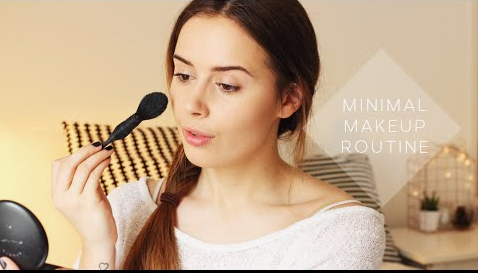 The Minimal Makeup Guide: How to Look Polished With Less Makeup A woman holding a makeup brush and looking into a mirror for a minimal makeup routine guide.