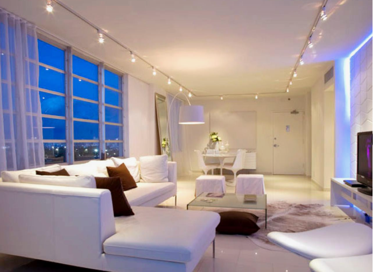 A contemporary, bright apartment living room featuring a white sectional sofa, track lighting, glowing blue LED wall accents, and large windows looking out at dusk.
