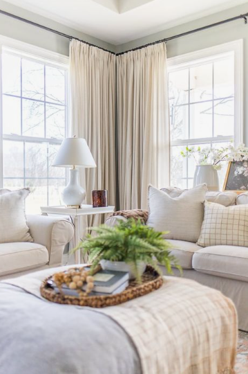 A cozy, bright living room corner featuring neutral-toned floor-to-ceiling drapes hanging from a dark rod between two large windows, a comfortable sofa, and an ottoman styled with a tray and greenery.