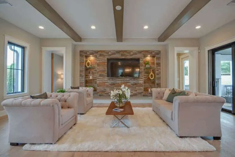 A spacious, modern living room featuring exposed ceiling beams, a stacked stone accent wall with a mounted television and built-in niches, a plush white rug, and classic beige tufted sofas.