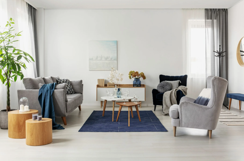A stylish, budget-friendly living room featuring a grey sofa with a blue throw blanket, a wingback armchair, round wooden side tables, a central blue rug, and minimalist artwork, demonstrating how to make a home look expensive on a budget.