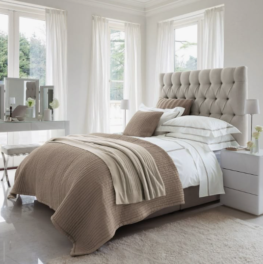 A bright, elegant modern bedroom featuring a tall tufted upholstered headboard, crisp white and beige layered bedding, sheer curtains framing the windows, and a sleek bedside table.