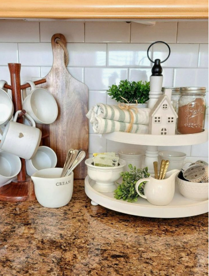 A cozy, organized kitchen counter featuring a wooden mug tree, a tall wooden cutting board, and a two-tiered white tray styled with greenery, jars, and decorative items to look expensive on a budget.