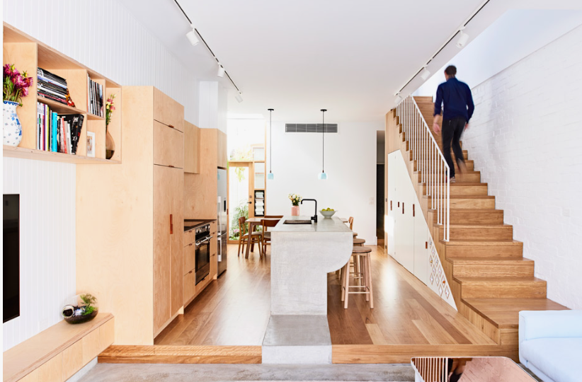 modern, open-plan apartment interior featuring a streamlined kitchen island, built-in light wood cabinetry and shelving, a staircase with a person walking up, and hardwood floors styled with clean lines for modern apartment living room ideas.