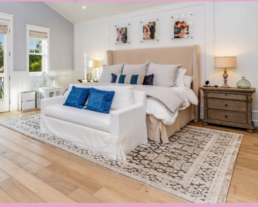 A bright, luxurious modern bedroom featuring a large beige tufted headboard, a white bench at the foot of the bed with royal blue accent pillows, a patterned area rug over light hardwood floors, and framed artwork mounted on a wainscotted wall.