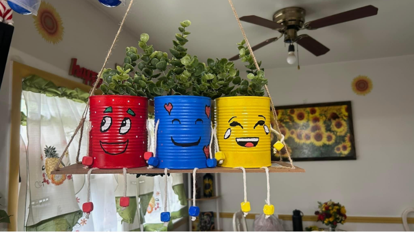 A hanging shelf holding three brightly colored tin cans with painted facial expressions, serving as planters for a green plant.