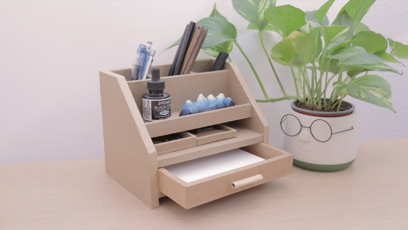 A beige DIY desk organizer crafted from cardboard, featuring upper pen compartments, open shelves, and a pull-out bottom drawer.