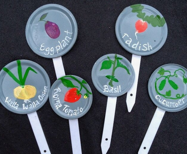 A collection of DIY garden markers made from painted jar lids attached to white stakes, featuring illustrations and labels for eggplant, radish, Walla Walla onion, Roma tomato, basil, and cucumber.