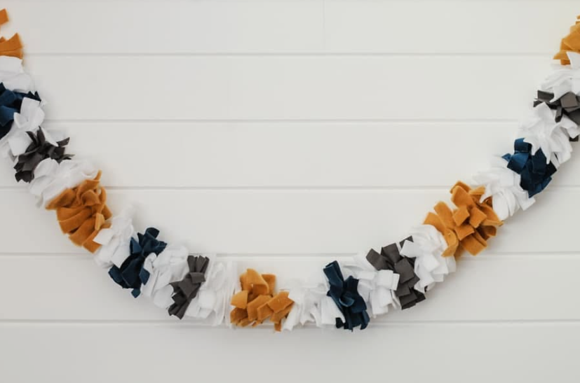 A fabric rag garland crafted from tied strips of white, dark blue, gray, and mustard yellow fabric, hanging against a white paneled wall.