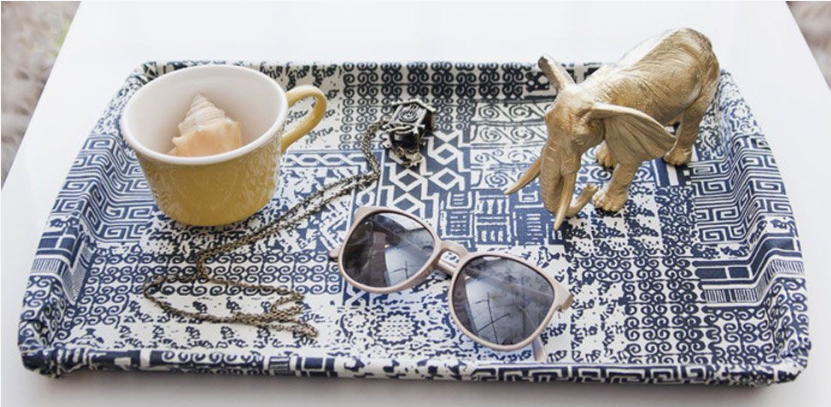 A blue and white patterned tray styled with a yellow cup containing a seashell, a gold elephant figurine, sunglasses, and a necklace.