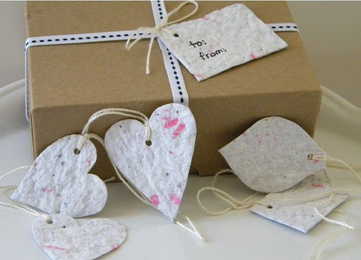 Handmade seed paper gift tags and heart-shaped cutouts featuring textured, recycled paper embedded with tiny specks and pink petals, tied with twine next to a wrapped cardboard box.