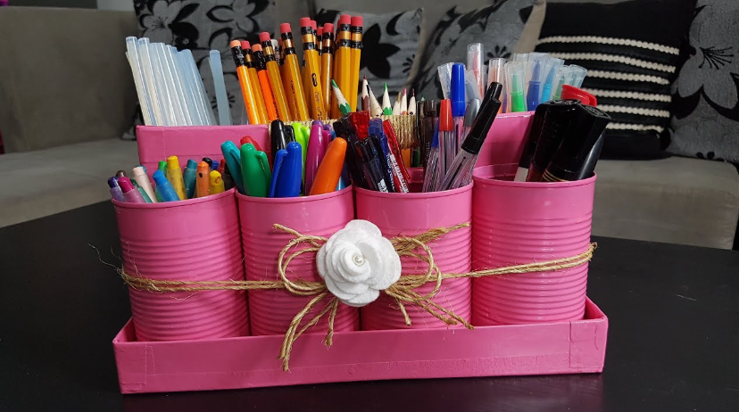 A DIY desk organizer crafted from painted pink tin cans mounted on a matching tray, decorated with twine and a white fabric flower, holding various pens, pencils, and glue sticks.