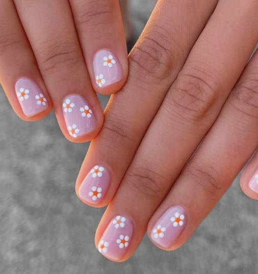 Short, rounded nails painted with a soft, translucent pink base coat and decorated with playful white daisy patterns featuring bright orange centers, capturing simple and easy nail designs for short nails.