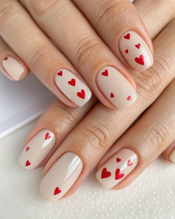 Short, glossy nude-beige nails decorated with scattered tiny red heart accents of varying sizes, serving as a charming example of easy nail designs for short nails.