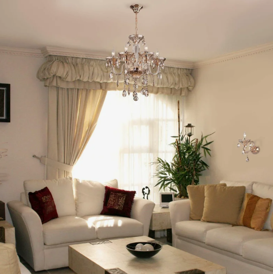 A classic living room featuring cream-colored sofas with accent pillows, a wooden coffee table, an ornate crystal chandelier hanging from the ceiling, layered window drapes, and a matching wall sconce, offering timeless design inspiration for renter-friendly home decor ideas.