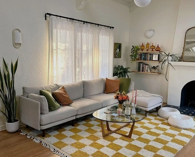 A cozy living room featuring a light gray sectional sofa styled with colorful throw pillows, an oval glass-top coffee table on a yellow and white checkered rug, wall-mounted wooden shelves filled with books and decor, and lush green house plants, offering charming inspiration for renter-friendly home decor ideas.