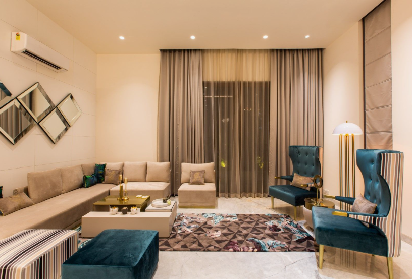 A modern living room featuring a long beige sectional sofa with accent pillows, a matching single seat, a decorative geometric mirror arrangement on the wall, teal velvet armchairs, a patterned area rug, floor-to-ceiling drapes, and a tall gold floor lamp, offering elegant design inspiration for renter-friendly home decor ideas.