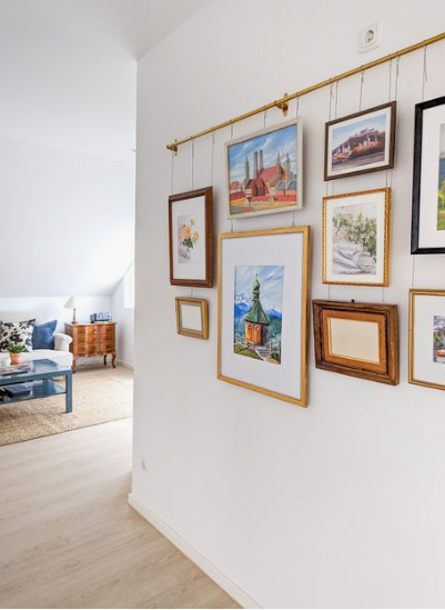 A bright hallway wall featuring an innovative brass picture hanging rail system with various framed artworks suspended by wires, providing renter-friendly home decor ideas for displaying a gallery wall without putting holes in the walls.