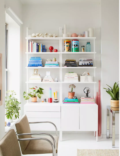 A bright room featuring modern white modular shelving units filled with colorful books, decorative objects, and small plants, paired with metal-framed armchairs and a small side table, offering stylish inspiration for renter-friendly home decor ideas.
