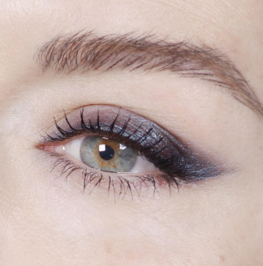 A macro beauty shot highlighting Spring 2026 makeup trends, featuring a feathery brow, smoky smudged charcoal eyeliner, and a green-blue hazel eye.