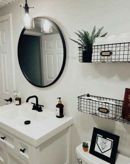 A modern bathroom featuring a white vanity with a black faucet, a round black-framed mirror with a matching pendant light above, and black wire wall baskets holding rolled towels and toilet paper, offering clever organization inspiration for renter-friendly home decor ideas.