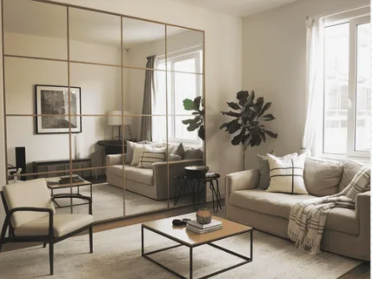 A chic and airy living room featuring a large floor-to-ceiling grid mirror wall that visually expands the space, paired with a beige sofa with patterned pillows, a square wood-top coffee table, an accent armchair, and a potted house plant near the window, offering clever design inspiration for renter-friendly home decor ideas.