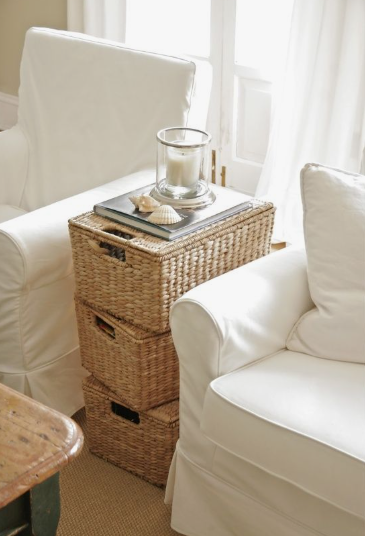 A clever space-saving setup featuring a stack of woven wicker storage baskets used as a casual side table between two white armchairs, styled with a tray, a glass-encased candle, and seashells, offering rustic and practical inspiration for renter-friendly home decor ideas.