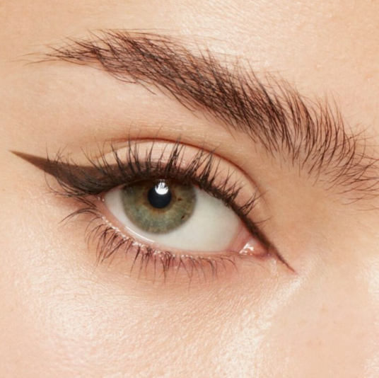 A macro beauty shot highlighting Spring 2026 makeup trends, featuring brushed-up feathery brows, sharp winged eyeliner, and a green iris.