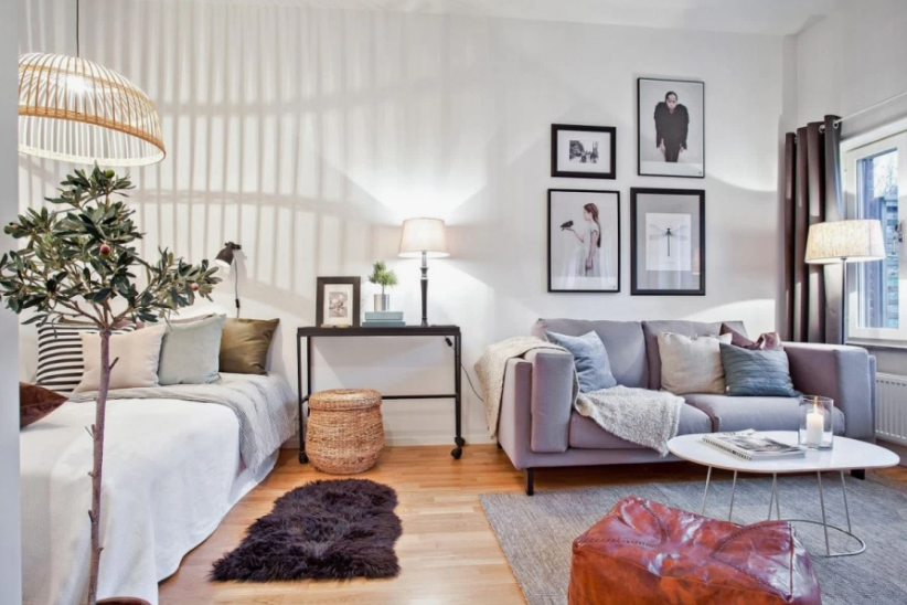 A cozy multi-functional living and sleeping space featuring a daybed with neutral bedding, a grey sofa with throw pillows and a knitted blanket, a gallery wall of framed black-and-white art, a white coffee table with a leather pouf, and a woven pendant light, offering creative inspiration for renter-friendly home decor ideas.