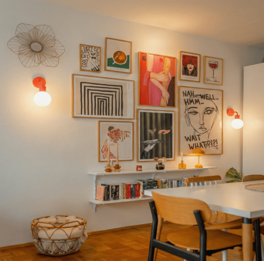 A vibrant gallery wall featuring an eclectic mix of framed art prints, a wire flower wall sculpture, a floating shelf holding books, and two glowing red-based wall sconces flanking the arrangement, offering creative inspiration for renter-friendly home decor ideas.