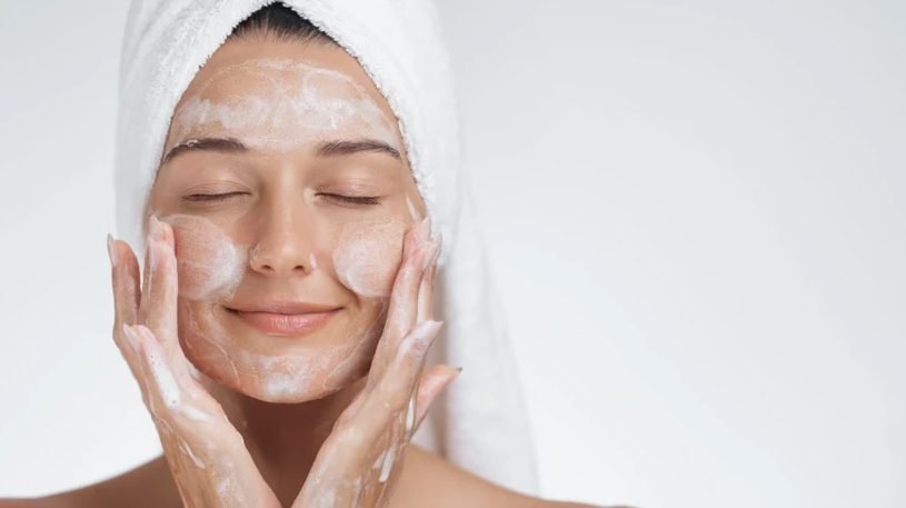 A close-up of a smiling woman with a white towel wrapped around her hair as she gently washes her face with a foaming cleanser, offering practical inspiration for a simple skincare routine for beginners.