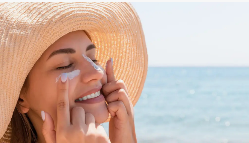 A smiling woman wearing a wide-brimmed straw hat at the beach as she applies sunscreen to her face, offering practical inspiration for a simple skincare routine for beginners.