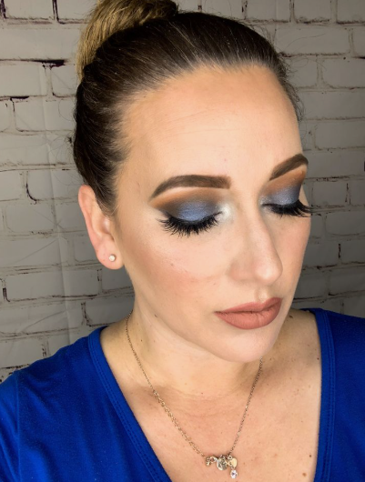 A beauty portrait highlighting Spring 2026 makeup trends, featuring a model wearing a royal blue top, bold smoky blue eyeshadow, defined eyebrows, and matte nude lips.