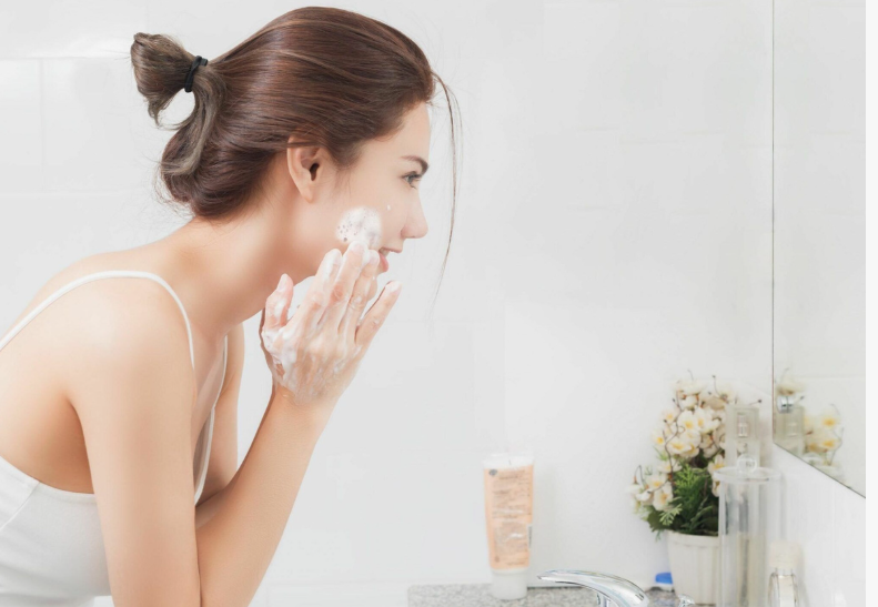A side-profile view of a woman with brown hair tied back in a bun as she gently lathers a foaming cleanser onto her cheek in front of a bathroom mirror, offering practical instruction for a simple skincare routine for beginners.