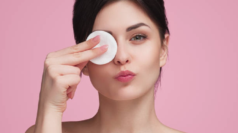 A close-up of a smiling woman gently removing makeup with a white cotton pad over her eye against a soft pink background, offering useful tips for a simple skincare routine for beginners.