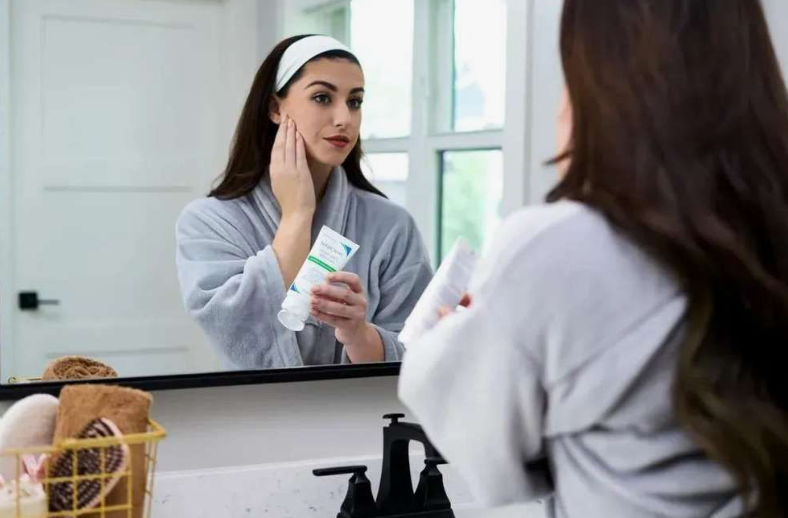 A woman wearing a gray bathrobe and a white headband checks her reflection in a bathroom mirror while applying lotion from a tube, offering a helpful visual for a simple skincare routine for beginners.