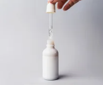 A hand holding a glass dropper filled with clear liquid over a white cosmetic serum bottle against a clean neutral background, offering a practical visual for a simple skincare routine for beginners.