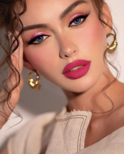 A close-up portrait of a model showcasing Spring 2026 makeup trends, featuring shimmering pink eyeshadow, sharp cat-eye liner, flushed cheeks, and vibrant magenta lips.