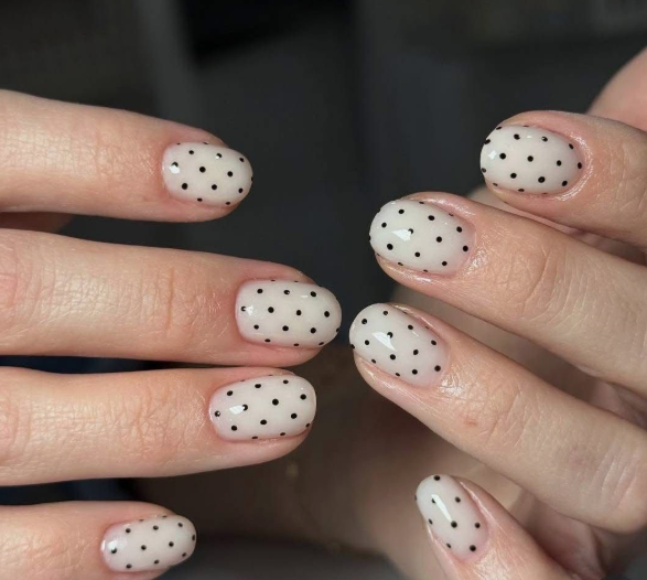 Short, rounded nails with a glossy milky white base featuring playful black polka dots, acting as a stylish choice for nail designs for short nail beds.