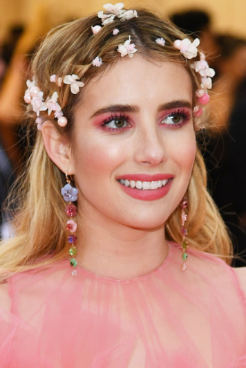 A beauty portrait highlighting Spring 2026 makeup trends, featuring a model wearing a pink dress adorned with flower hair accessories, monochromatic pink eyeshadow, rosy cheeks, and pink lips.
