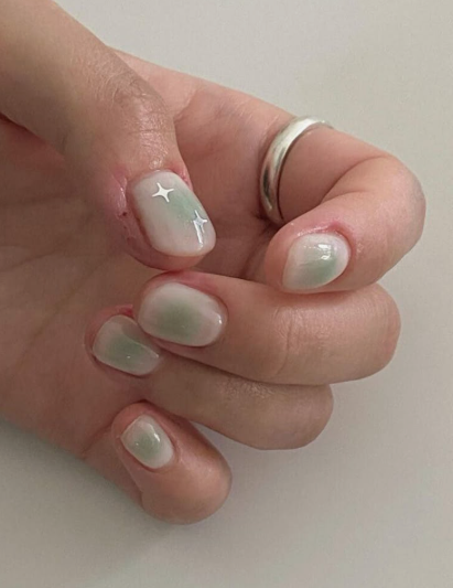 Short, rounded nails featuring a soft milky ombre base with a diffused sage green aura effect and delicate silver star decals, making it a dreamy option for nail designs for short nail beds.
