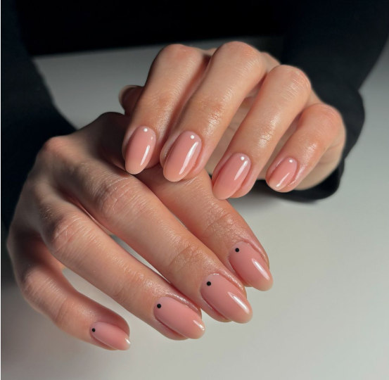 Short, rounded nails with a glossy peach-pink nude polish featuring a single tiny black dot accent at the base of each nail, creating a minimalist choice for nail designs for short nail beds.