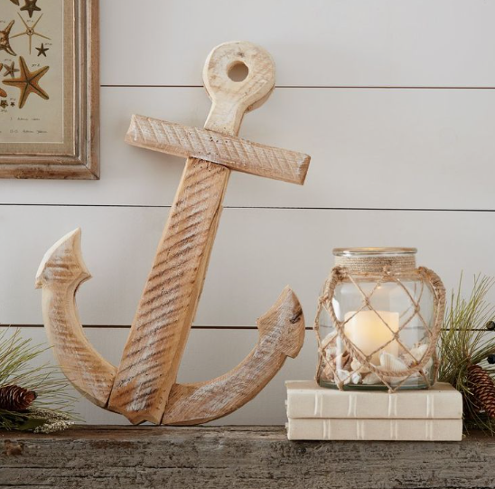 A rustic, weathered wooden anchor decor piece displayed alongside a rope-wrapped glass candle jar resting on small books, perfectly embodying the keyphrase how to decorate when everything doesn't match by blending varied textures and coastal elements.